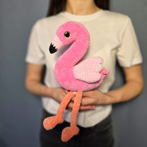 May include: A pink crocheted flamingo toy with black eyes and a black beak. The flamingo is sitting on its legs with its wings spread out. The toy is being held by a person.