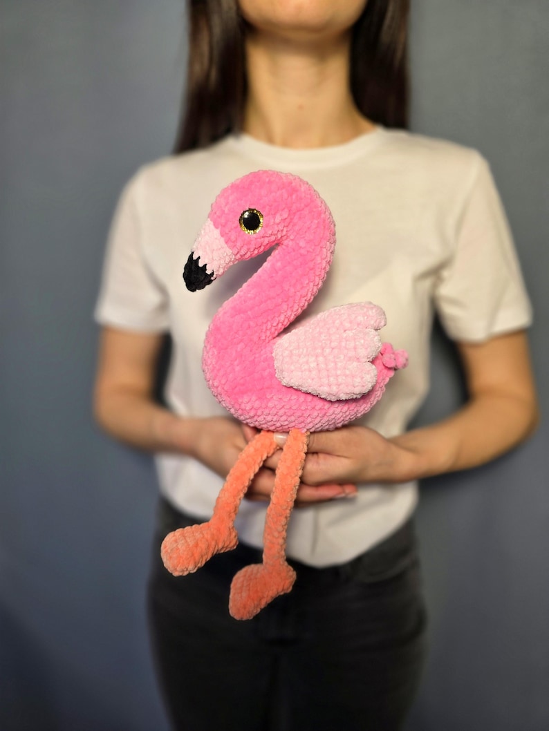 May include: A pink crocheted flamingo toy with black eyes and a black beak. The flamingo is sitting on its legs with its wings spread out. The toy is being held by a person.
