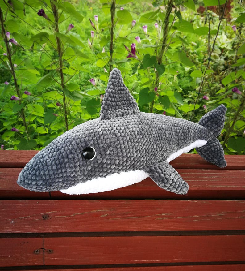 Op de afbeelding: Een grijs pluche haaien speelgoed met een witte buik en zwarte ogen. De haai ligt op een houten oppervlak.