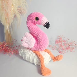 May include: Pink crocheted flamingo toy sitting on a ball of white yarn. The flamingo has black eyes and a black beak.