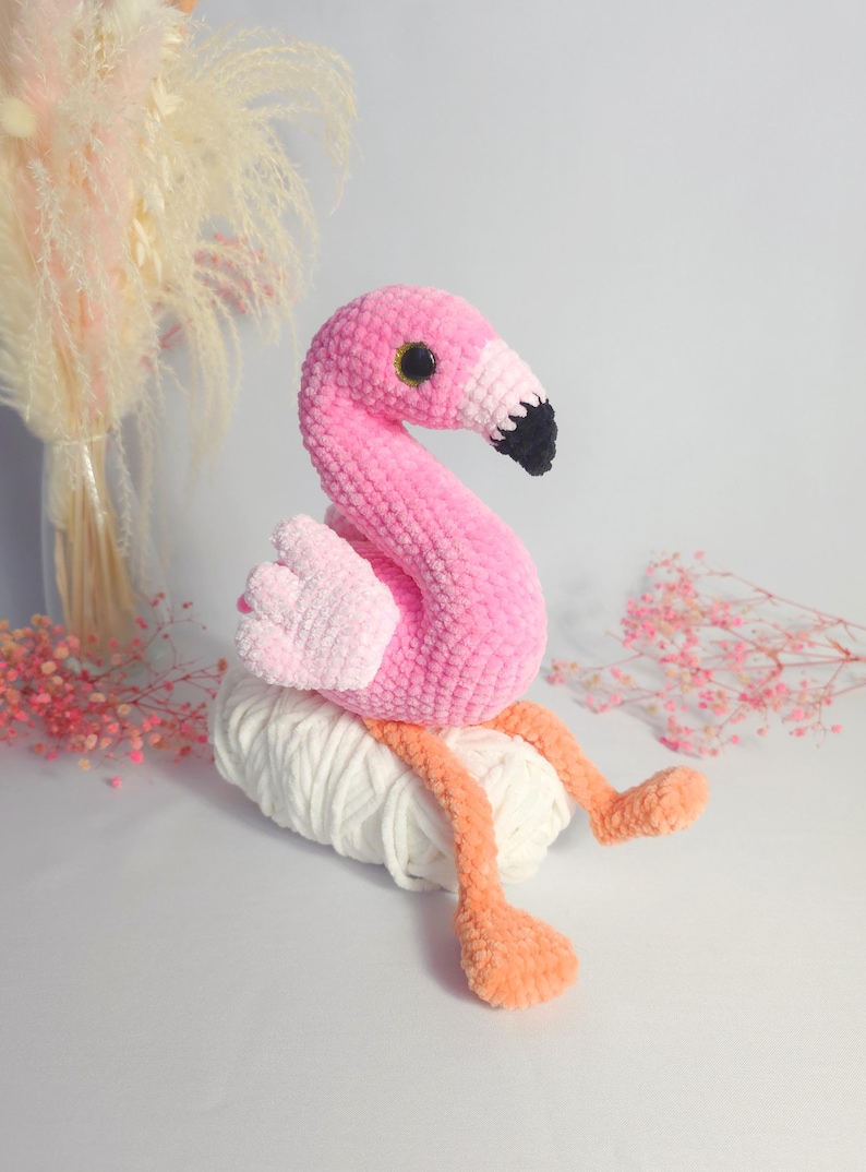 May include: Pink crocheted flamingo toy sitting on a ball of white yarn. The flamingo has black eyes and a black beak.