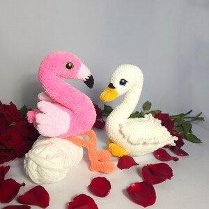 以下が含まれることがあります： 刺繍された目のついたピンクのフラミンゴと白い白鳥のぬいぐるみが、赤いバラの花びらが散りばめられた白い表面に座っています。