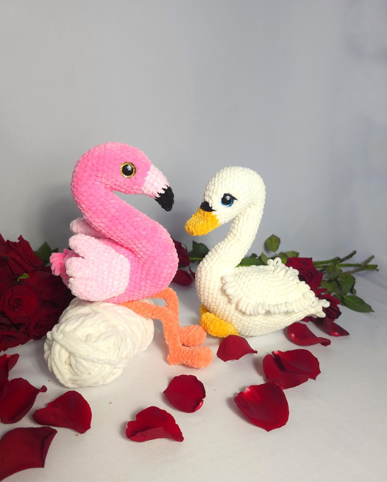 以下が含まれることがあります： 刺繍された目のついたピンクのフラミンゴと白い白鳥のぬいぐるみが、赤いバラの花びらが散りばめられた白い表面に座っています。