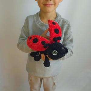 May include: A child holds a crocheted ladybug toy with red wings and black spots. The ladybug has black yarn for its body and legs, and large black eyes.