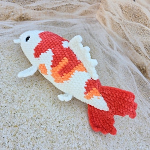 Pu&ograve; includere: Un giocattolo di peluche all'uncinetto a forma di pesce koi. Il pesce &egrave; bianco con accenti arancioni e rossi. Il pesce &egrave; sdraiato su una superficie sabbiosa.