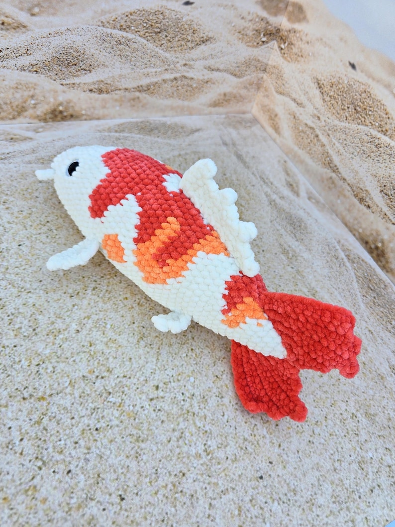 Pu&ograve; includere: Un giocattolo di peluche all'uncinetto a forma di pesce koi. Il pesce &egrave; bianco con accenti arancioni e rossi. Il pesce &egrave; sdraiato su una superficie sabbiosa.