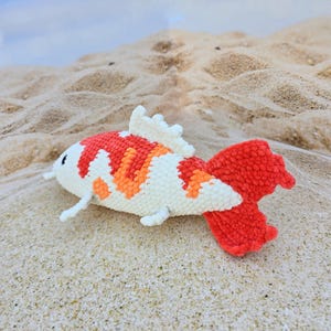 Pu&ograve; includere: Un giocattolo di peluche all'uncinetto a forma di pesce koi. Il pesce &egrave; bianco con accenti arancioni e rossi. Il pesce &egrave; sdraiato su una superficie sabbiosa.