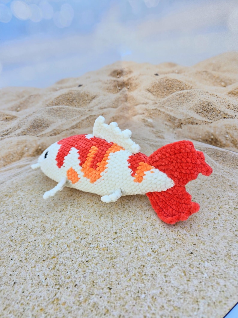 Pu&ograve; includere: Un giocattolo di peluche all'uncinetto a forma di pesce koi. Il pesce &egrave; bianco con accenti arancioni e rossi. Il pesce &egrave; sdraiato su una superficie sabbiosa.