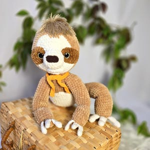 May include: A crocheted sloth plush toy with brown fur, white accents, and a yellow scarf. The sloth is sitting on a woven wicker basket.