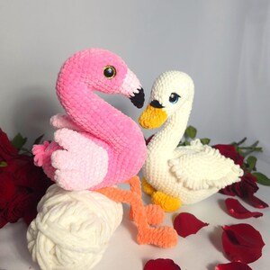 以下が含まれることがあります： ピンクのフラミンゴと白い白鳥のぬいぐるみは、どちらも編み物でできており、白い毛糸の玉の上に座っています。フラミンゴは長いオレンジ色の首と脚を持っています。白鳥は黄色いくちばしと青い目を持っています。おもちゃは赤いバラの花びらで囲まれています。
