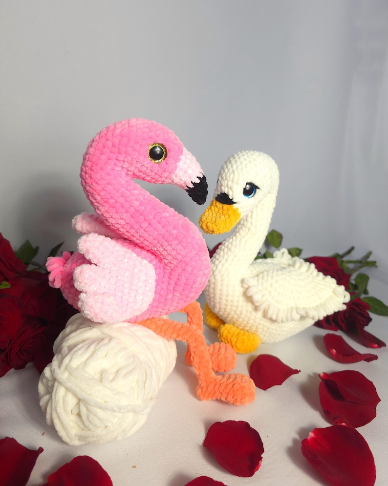 以下が含まれることがあります： ピンクのフラミンゴと白い白鳥のぬいぐるみは、どちらも編み物でできており、白い毛糸の玉の上に座っています。フラミンゴは長いオレンジ色の首と脚を持っています。白鳥は黄色いくちばしと青い目を持っています。おもちゃは赤いバラの花びらで囲まれています。