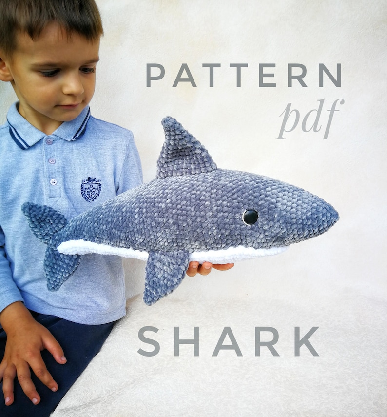 Op de afbeelding: Een grijs gehaakt haaien knuffel met witte accenten. Het speelgoed wordt vastgehouden door een kind. De tekst "PATTERN pdf" en "SHARK" is afgedrukt onder het speelgoed.