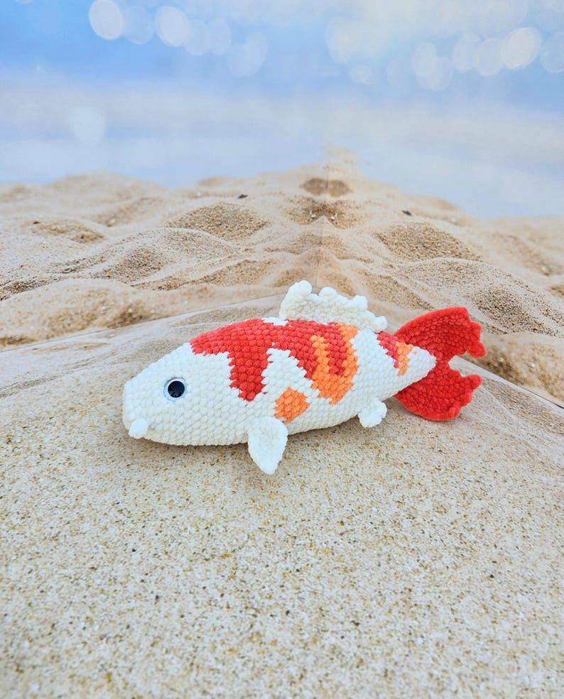 Pu&ograve; includere: Un giocattolo di pesce koi all'uncinetto con colori bianco, arancione e rosso. Il pesce &egrave; sdraiato su una superficie sabbiosa.