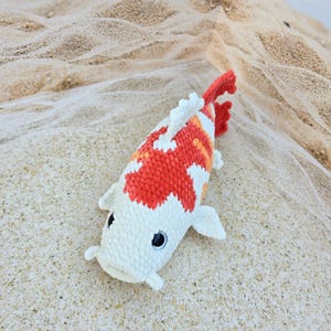 Pu&ograve; includere: Un giocattolo di peluche a forma di pesce koi in crochet bianco e arancione con occhi neri. Il giocattolo &egrave; sdraiato su una superficie sabbiosa.