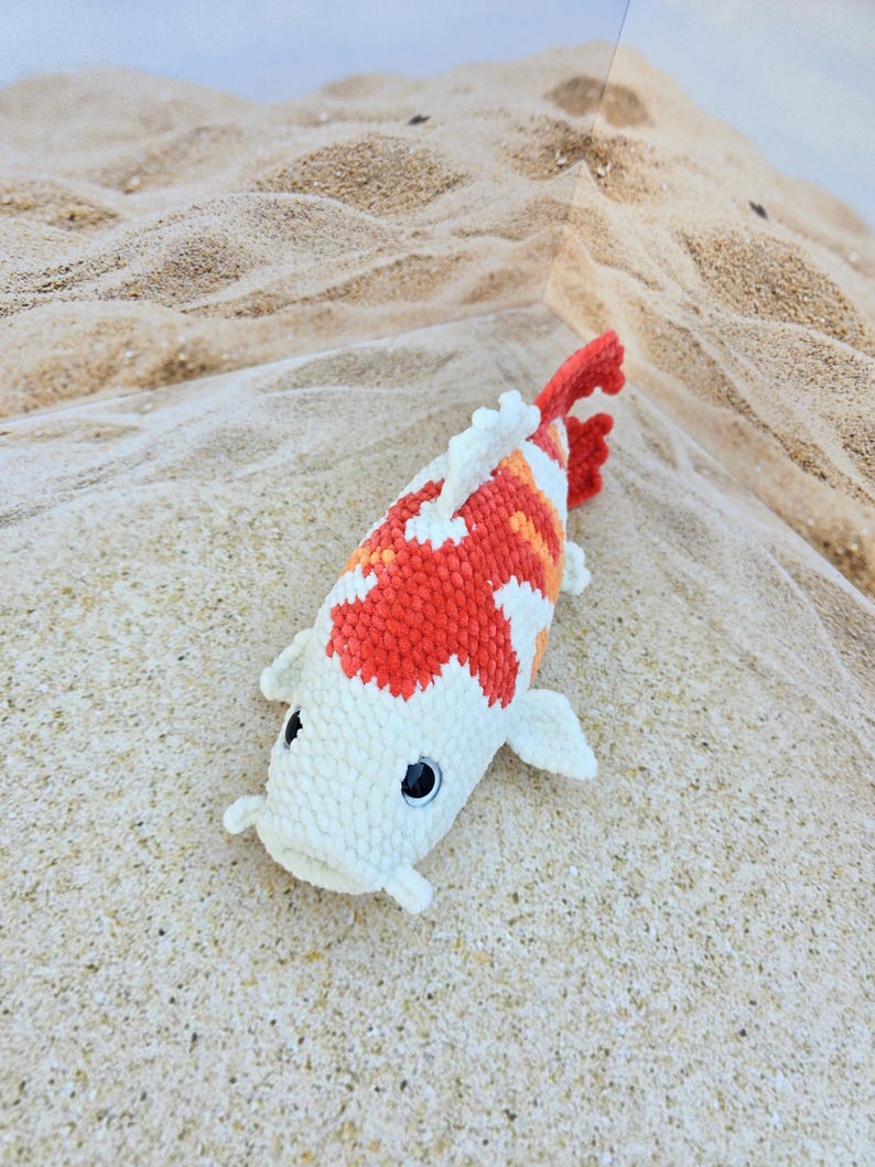 Pu&ograve; includere: Un giocattolo di peluche a forma di pesce koi in crochet bianco e arancione con occhi neri. Il giocattolo &egrave; sdraiato su una superficie sabbiosa.