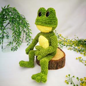 May include: A green crocheted frog plush toy with yellow accents and large black eyes. The frog is sitting on a wooden log.