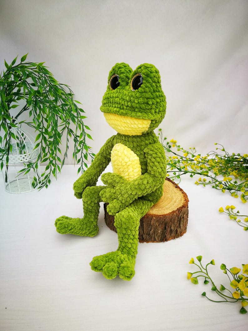 May include: A green crocheted frog plush toy with yellow accents and large black eyes. The frog is sitting on a wooden log.