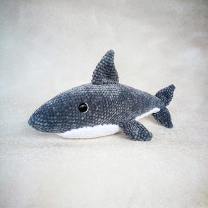 Op de afbeelding: Een grijs gehaakt knuffelhaai-speelgoed met witte details rond de mond. De haai heeft zwarte ogen en een textuur.