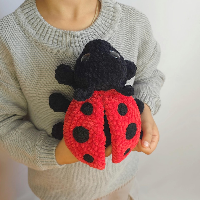 May include: A crocheted ladybug toy with a black head and red body with black spots. The ladybug is being held by a person wearing a gray sweater.