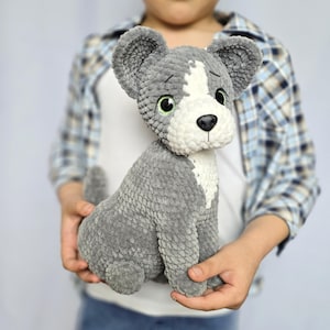 Puede incluir: Un juguete de peluche gris y blanco con forma de perro en posición sentada. El juguete tiene orejas grandes y caídas, una mancha blanca en la cara y el pecho, y ojos verdes. El perro es sostenido por una persona que lleva una camisa a cuadros.