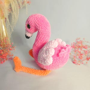 May include: A pink and orange crocheted flamingo toy. The flamingo is sitting on a white surface.