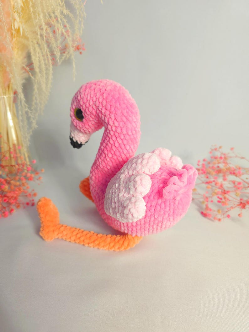 May include: A pink and orange crocheted flamingo toy. The flamingo is sitting on a white surface.