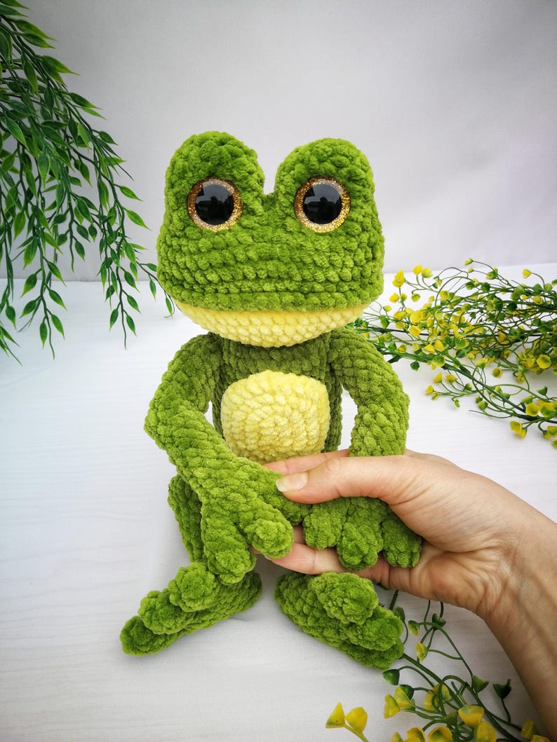 May include: Green crocheted frog plush toy with large, glittery eyes. The frog is sitting with its legs crossed and has a yellow belly.