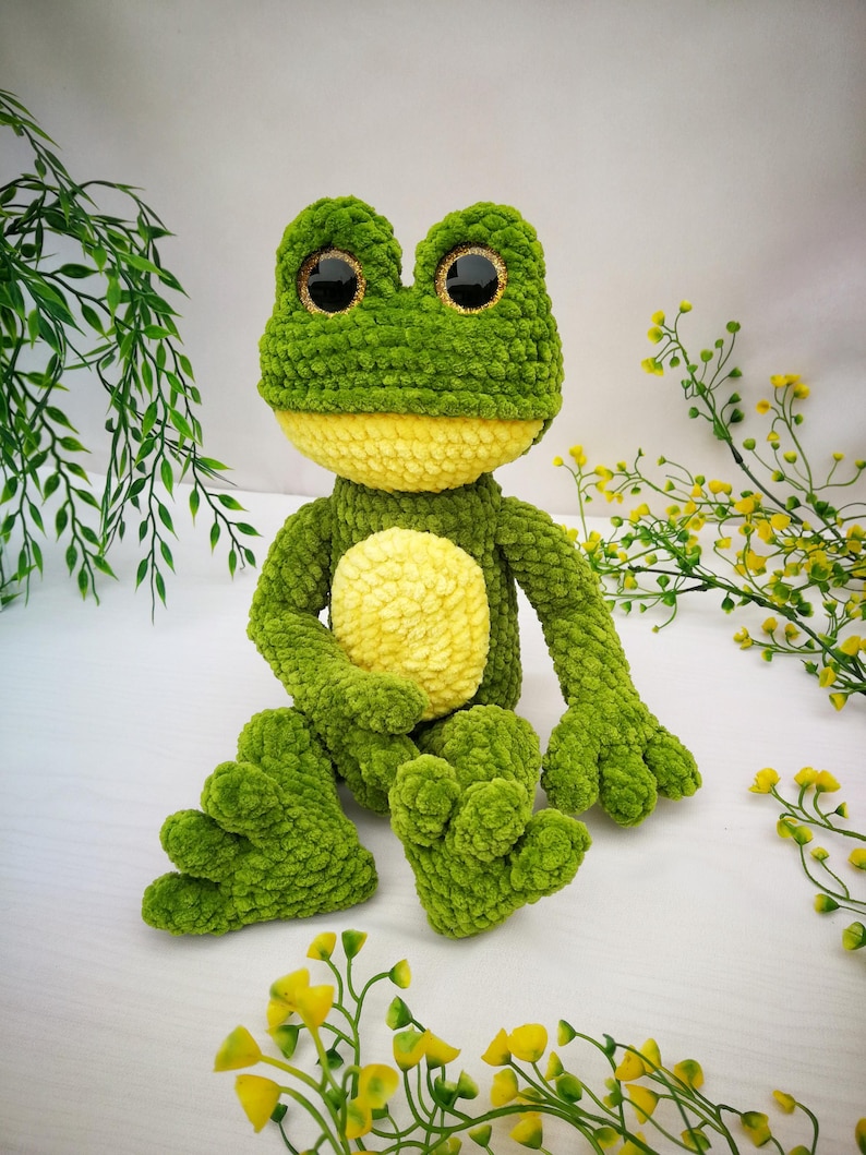 May include: A green crocheted frog plush toy with large, sparkly eyes. The frog is sitting with its legs crossed and its arms folded in front of it.