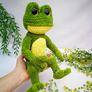 May include: A green crocheted frog plush toy with yellow accents and large black eyes. The frog is sitting on a hand.