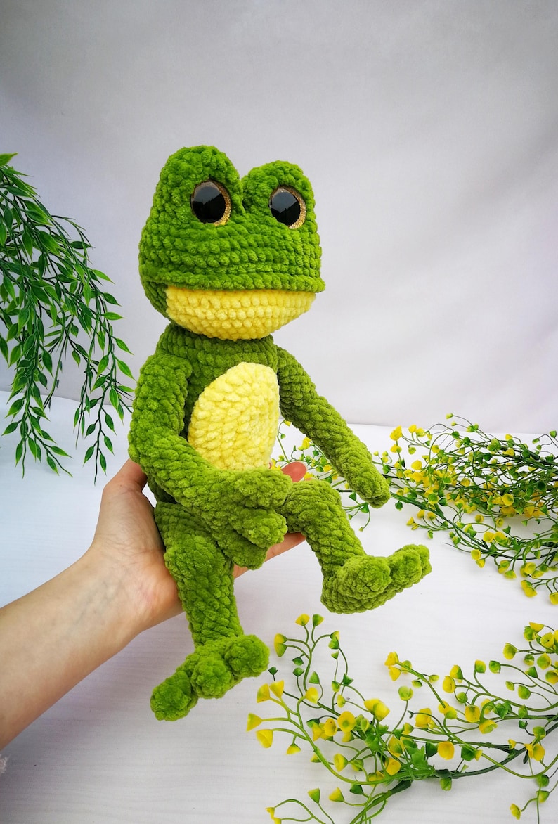 May include: A green crocheted frog plush toy with yellow accents and large black eyes. The frog is sitting on a hand.
