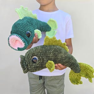 Könnte beinhalten: Zwei Plüschfisch-Spielzeuge in Grüntönen. Einer ist ein dunkleres Türkis mit einem rosa gefütterten Maul, der andere ist ein helleres Olivgrün. Beide haben große, runde Augen und hellgrüne Flossen. Die Spielzeuge sind weich und strukturiert.