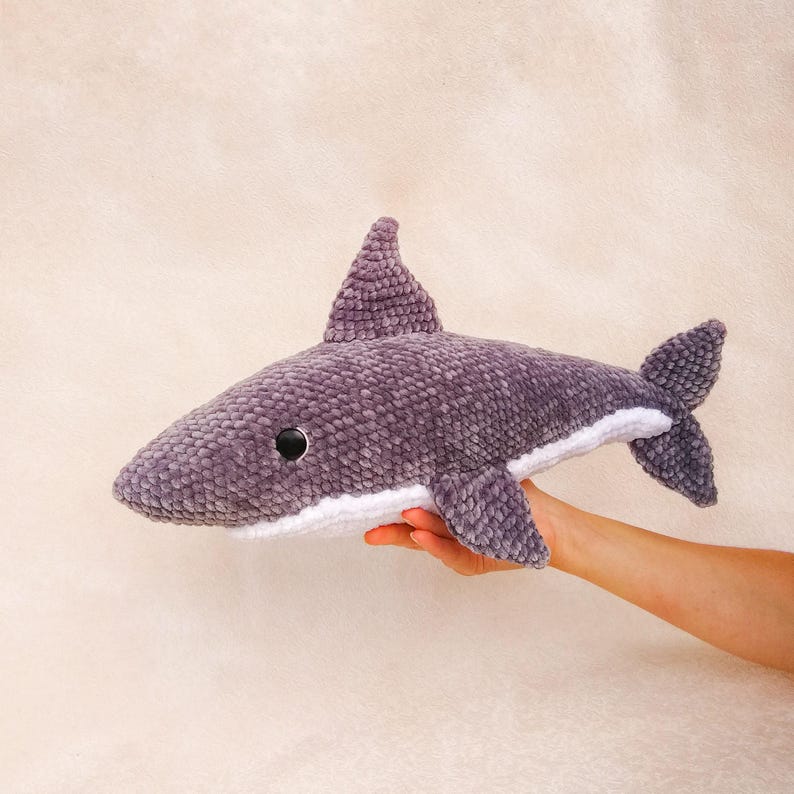 Op de afbeelding: Een grijs gehaakt knuffelhaai-speelgoed met witte details rond de mond. De haai heeft een zwart oog en wordt vastgehouden door een hand.