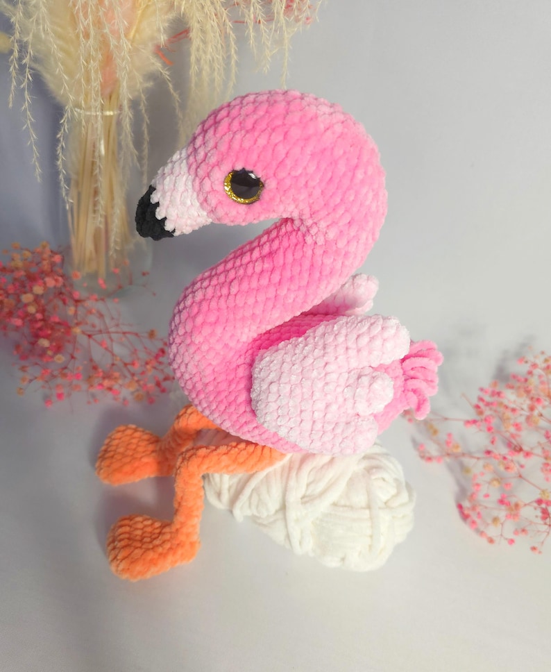 May include: A pink crocheted flamingo toy with orange legs sitting on a ball of white yarn. The flamingo has large black eyes and a pink and white ombre effect.