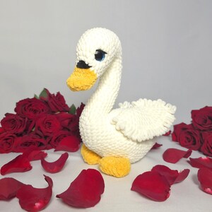 以下が含まれることがあります： 赤いバラの花びらに囲まれた、黄色いクチバシと青い目をした白い編みぐるみの白鳥