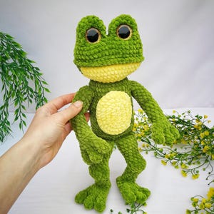 May include: A green crocheted frog plush toy with yellow accents and large black eyes. The frog is standing on a white surface with yellow flowers.