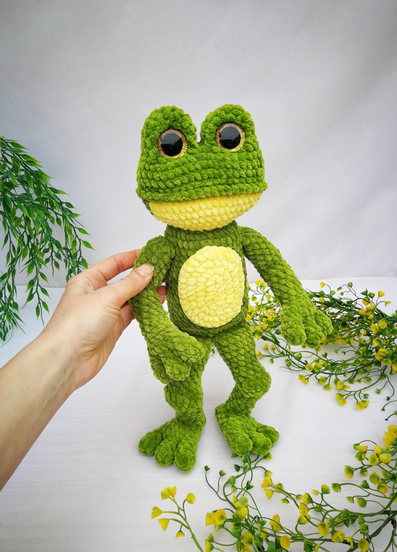May include: A green crocheted frog plush toy with yellow accents and large black eyes. The frog is standing on a white surface with yellow flowers.