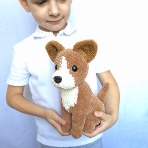 May include: A brown and white crocheted dog plush toy with large ears, a white chest, and a black nose. The handmade stuffed animal is held in a child's hands. The dog has a small tail and is made of soft, textured yarn.