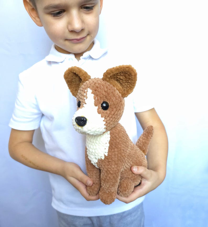 May include: A brown and white crocheted dog plush toy with large ears, a white chest, and a black nose. The handmade stuffed animal is held in a child's hands. The dog has a small tail and is made of soft, textured yarn.