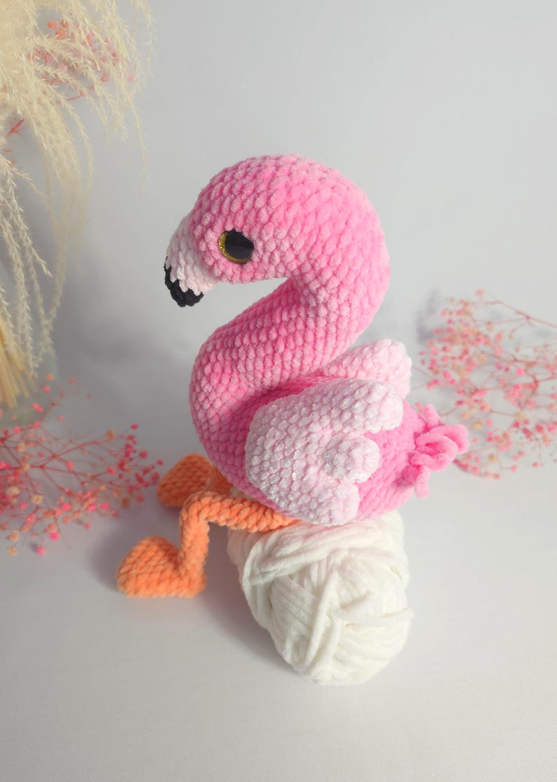 May include: A pink and white crocheted flamingo toy sitting on a ball of white yarn. The flamingo has black eyes and orange legs.