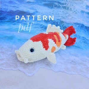 Pu&ograve; includere: Un giocattolo di peluche a forma di pesce koi all'uncinetto con squame bianche, arancioni e rosse. Il giocattolo &egrave; sdraiato su una spiaggia di sabbia bianca con acqua blu sullo sfondo. Il testo "PATTERN pdf" &egrave; visibile nell'angolo in alto a sinistra dell'immagine.