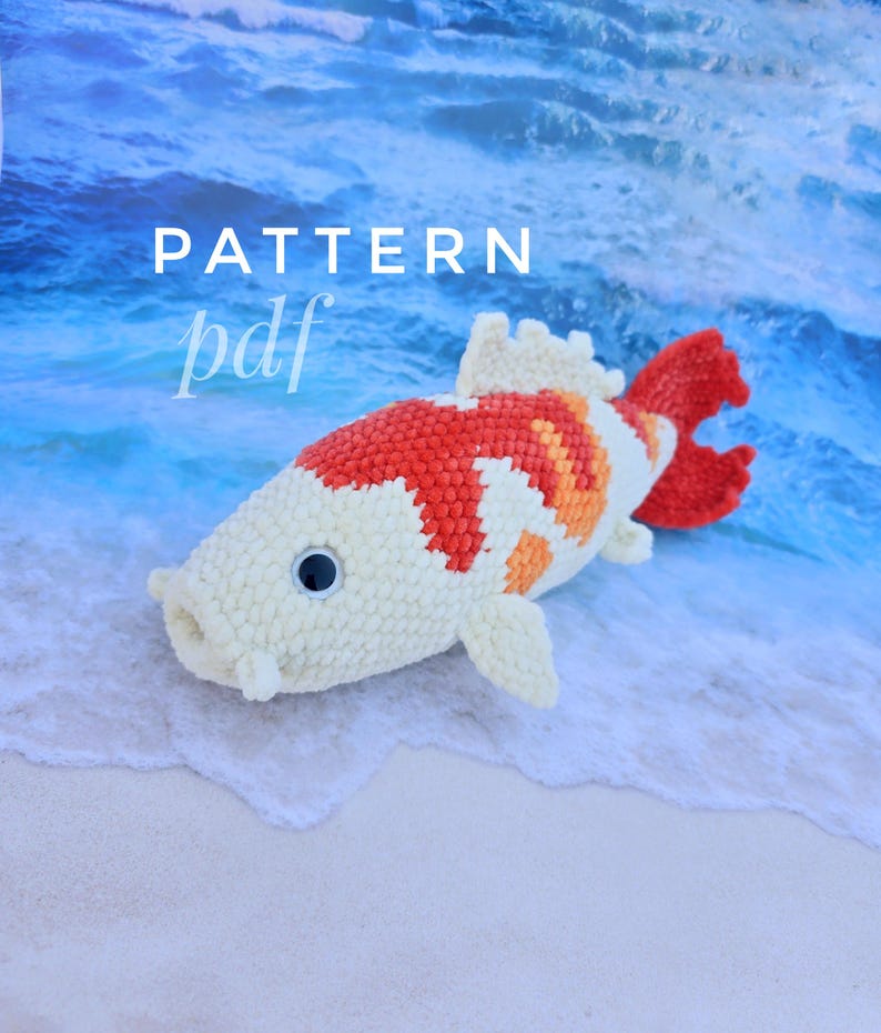 Pu&ograve; includere: Un giocattolo di peluche a forma di pesce koi all'uncinetto con squame bianche, arancioni e rosse. Il giocattolo &egrave; sdraiato su una spiaggia di sabbia bianca con acqua blu sullo sfondo. Il testo "PATTERN pdf" &egrave; visibile nell'angolo in alto a sinistra dell'immagine.