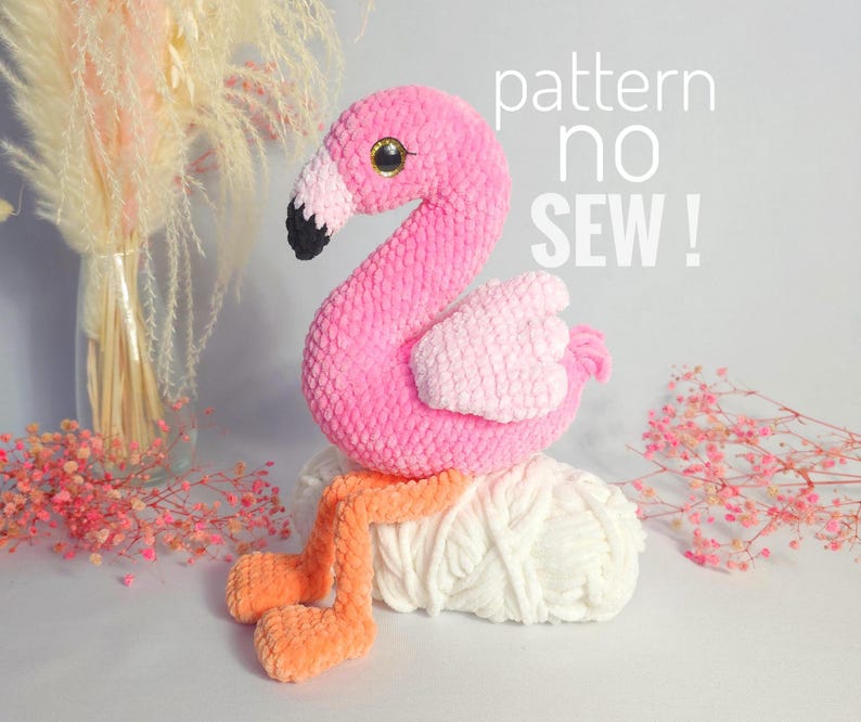 May include: Pink crocheted flamingo toy with orange legs. The flamingo is sitting on a white yarn ball. The text "pattern no sew!" is written in black on a white background.