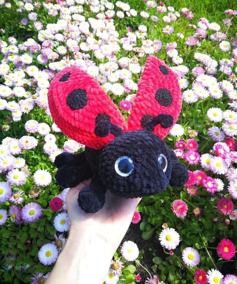 May include: A crocheted ladybug toy with black fur and red wings with black spots. The ladybug is being held in a hand against a background of white and pink flowers.