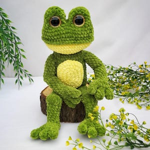 May include: A green crocheted frog plush toy with large black eyes and a yellow belly. The frog is sitting on a wooden log with green foliage in the background.