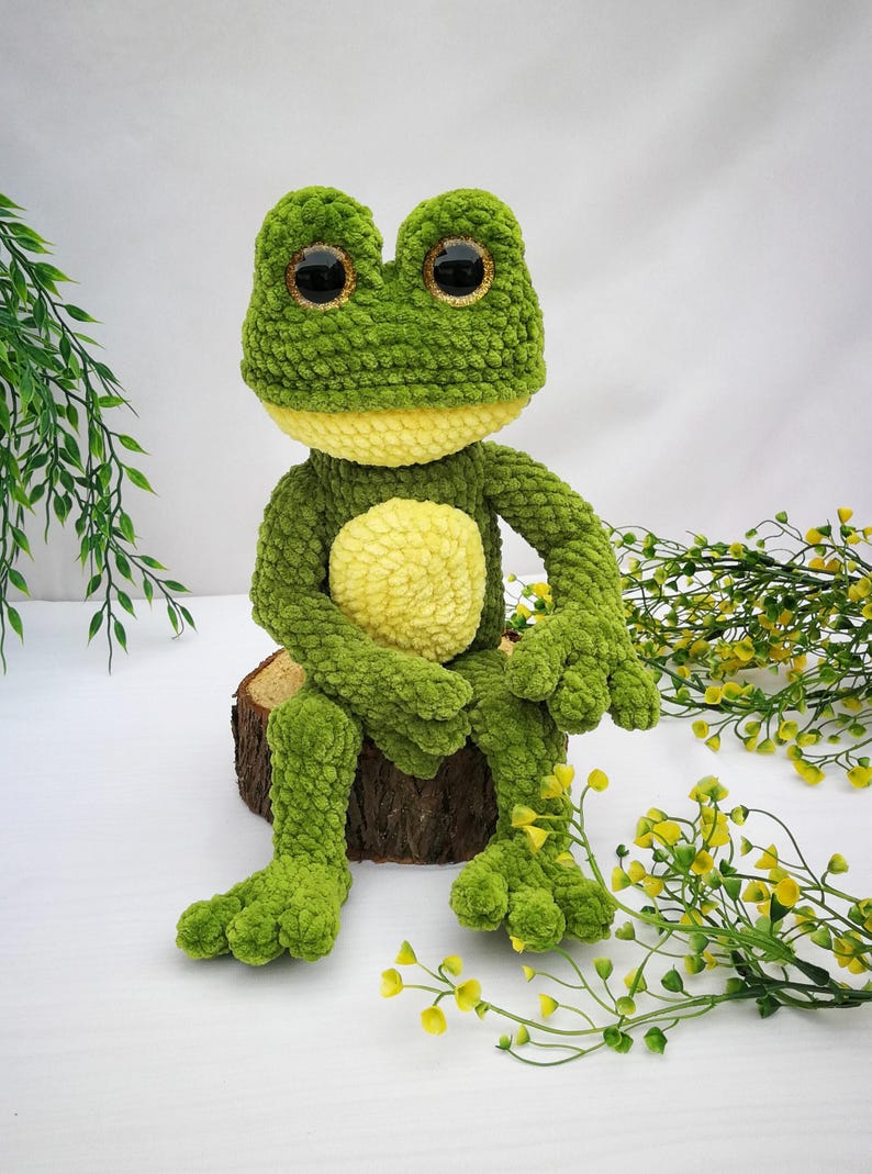 May include: A green crocheted frog plush toy with large black eyes and a yellow belly. The frog is sitting on a wooden log with green foliage in the background.