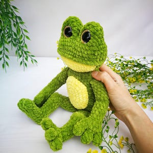 May include: Green crocheted frog plush toy with large black eyes and a yellow belly. The frog is sitting on a white surface with green and yellow flowers.