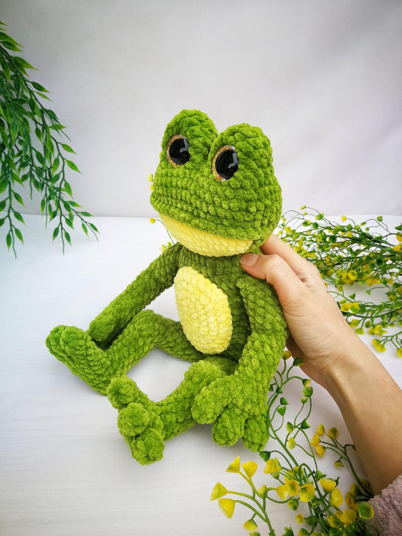 May include: Green crocheted frog plush toy with large black eyes and a yellow belly. The frog is sitting on a white surface with green and yellow flowers.