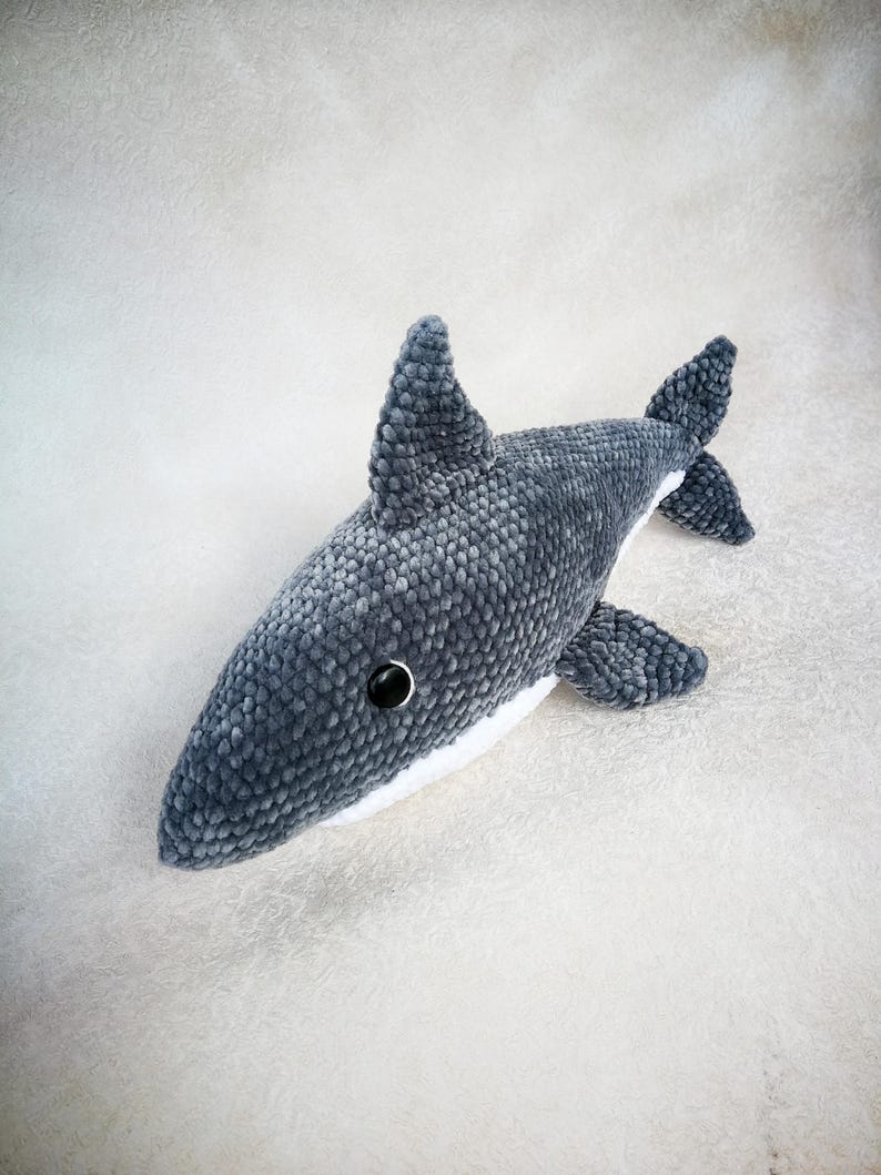 Op de afbeelding: Een grijs pluche haaien speelgoed met een witte buik en zwarte ogen. De haai ligt op een wit, pluche oppervlak.