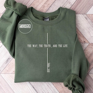 May include: An olive green sweatshirt with the embroidered word "EMBROIDERED" in a white circle. The front of the sweatshirt features a white cross design with the text "THE WAY, THE TRUTH, AND THE LIFE" and "John 14:6".
