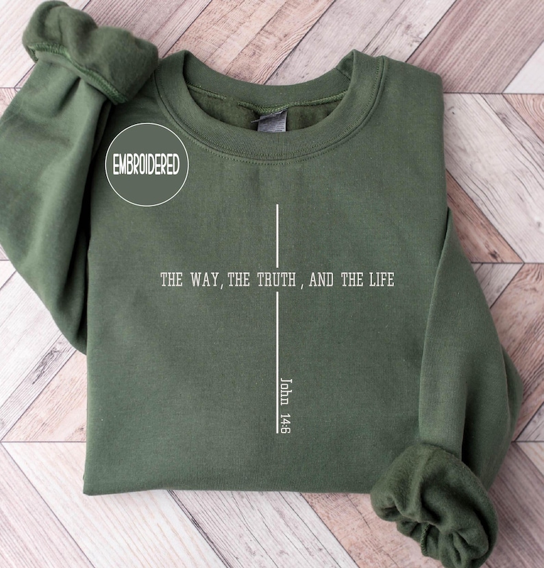 May include: An olive green sweatshirt with the embroidered word "EMBROIDERED" in a white circle. The front of the sweatshirt features a white cross design with the text "THE WAY, THE TRUTH, AND THE LIFE" and "John 14:6".